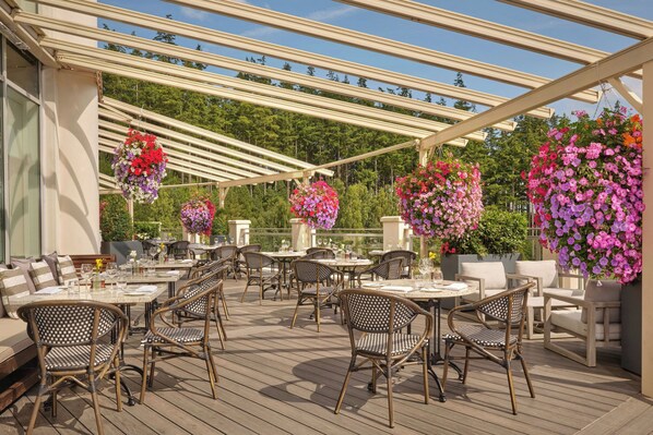 Un patio al aire libre con mesas y sillas, rodeado por una pérgola y cestas colgantes de flores.