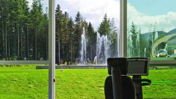 Ein Fitnessbereich mit Laufrad, Gymnastikbällen und Blick auf einen Brunnen sowie Bäume durch große Fenster.