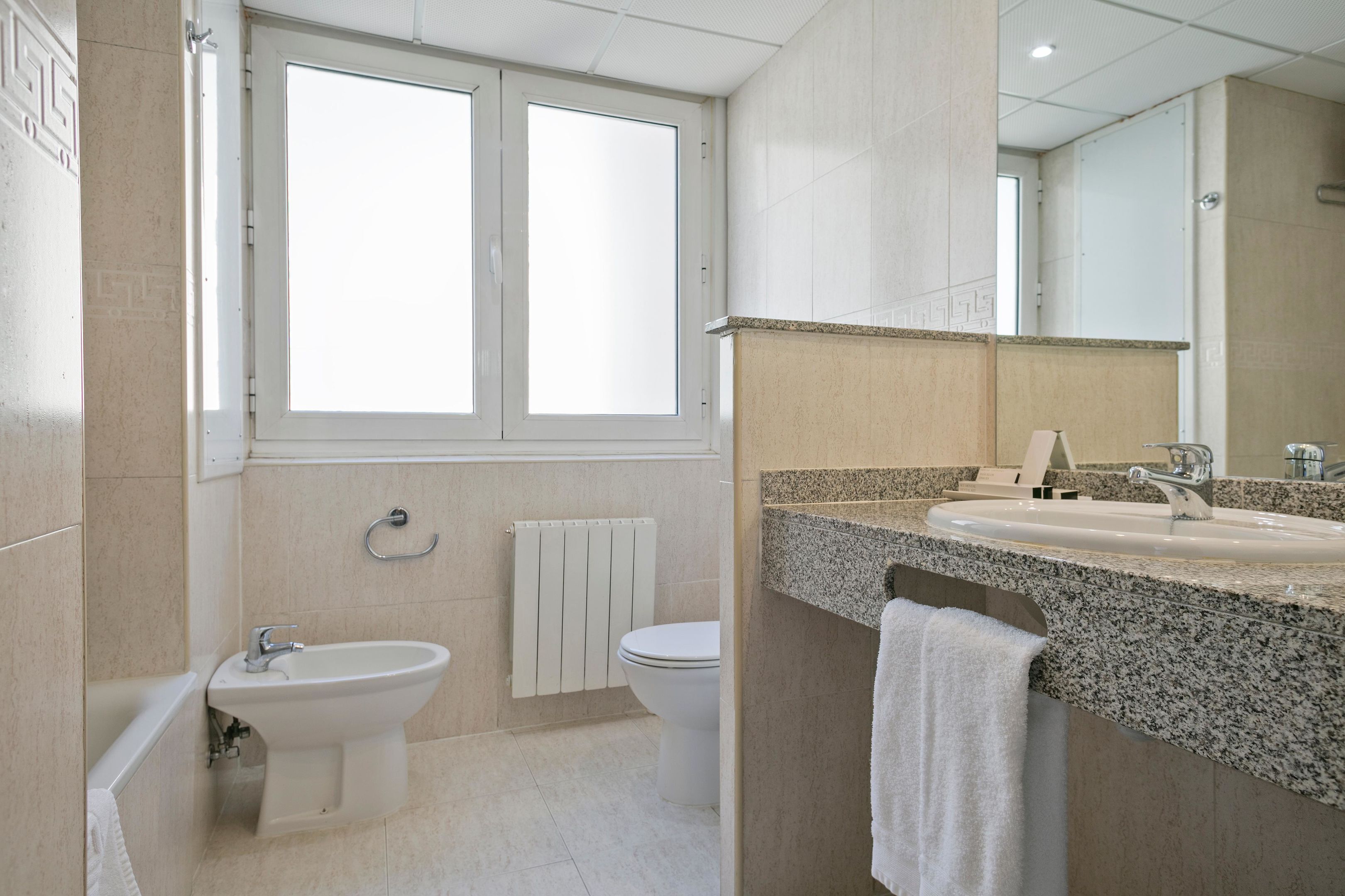 Un baño moderno con bidé, lavabo e inodoro, que cuenta con un espejo grande y una ventana.