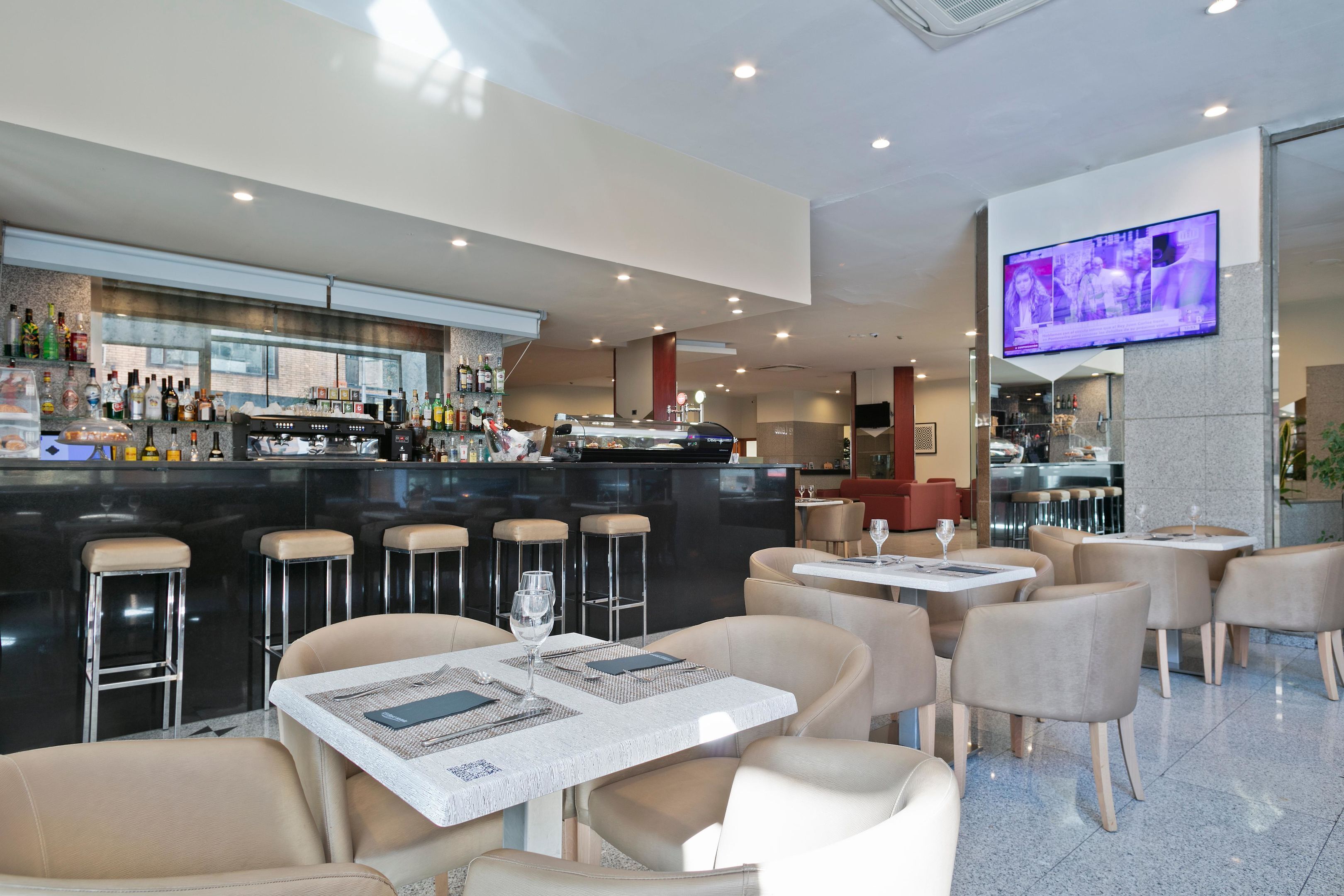 Een moderne bar met een lange toonbank, meerdere barkrukken en een televisie aan de muur.