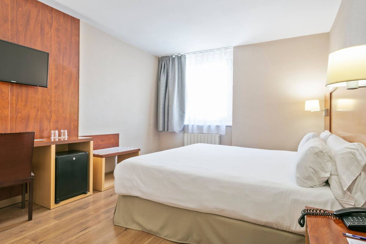 Una habitación de hotel con una cama, un escritorio con computadora, una silla, una lámpara y un teléfono.