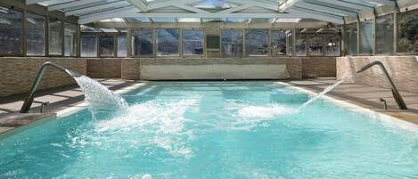 Una piscina con techo de vidrio, rodeada de ventanas y un muro de piedra.