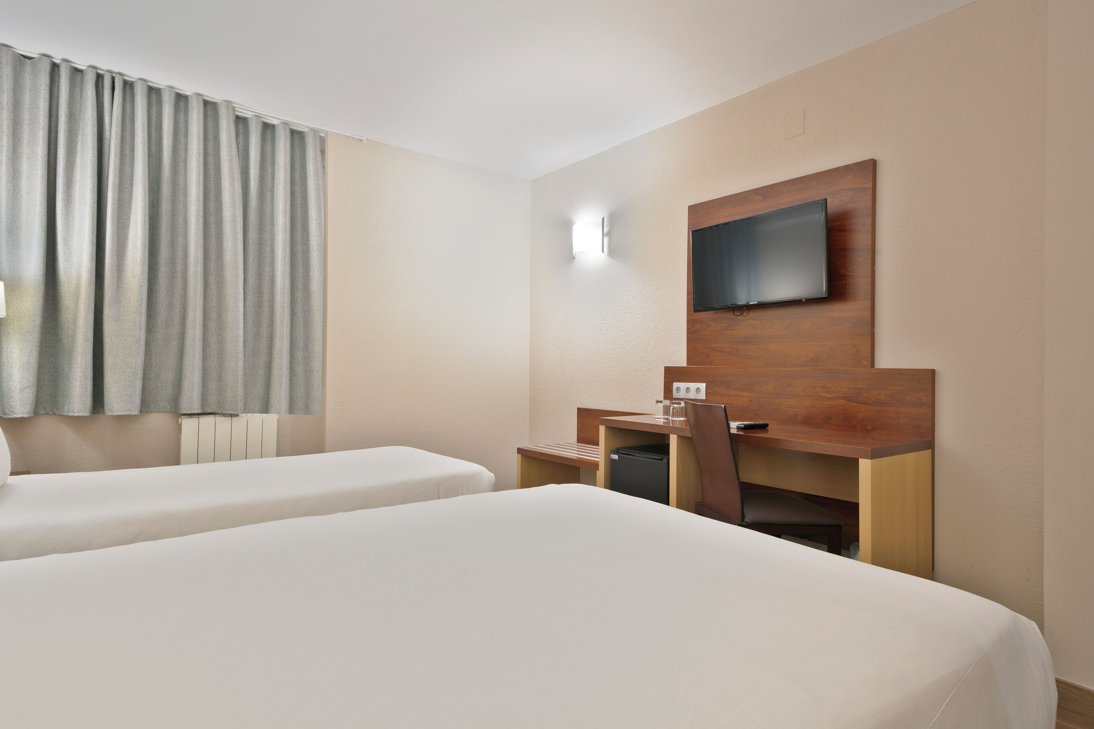 A hotel room with two beds, a desk, a chair, a TV mounted on the wall, and a window with curtains.