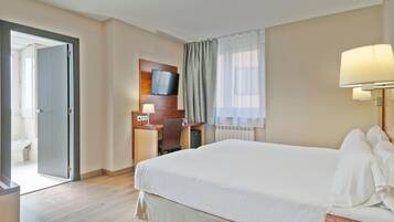 A hotel room with a bed, a desk with a chair, a television, and a window with curtains.