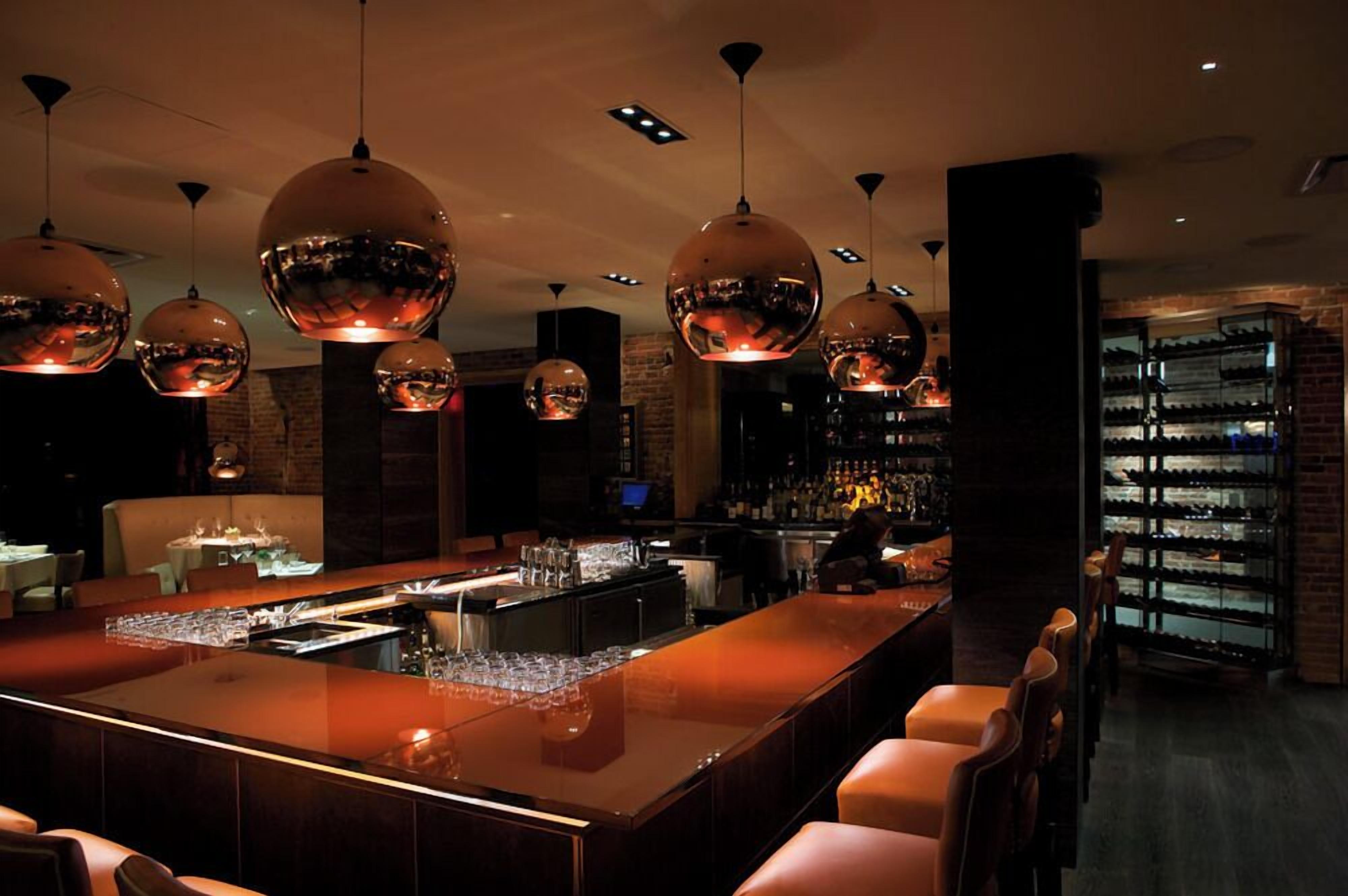 Zona de bar con una barra alargada, varias opciones de asientos y una estantería de vinos al fondo.
