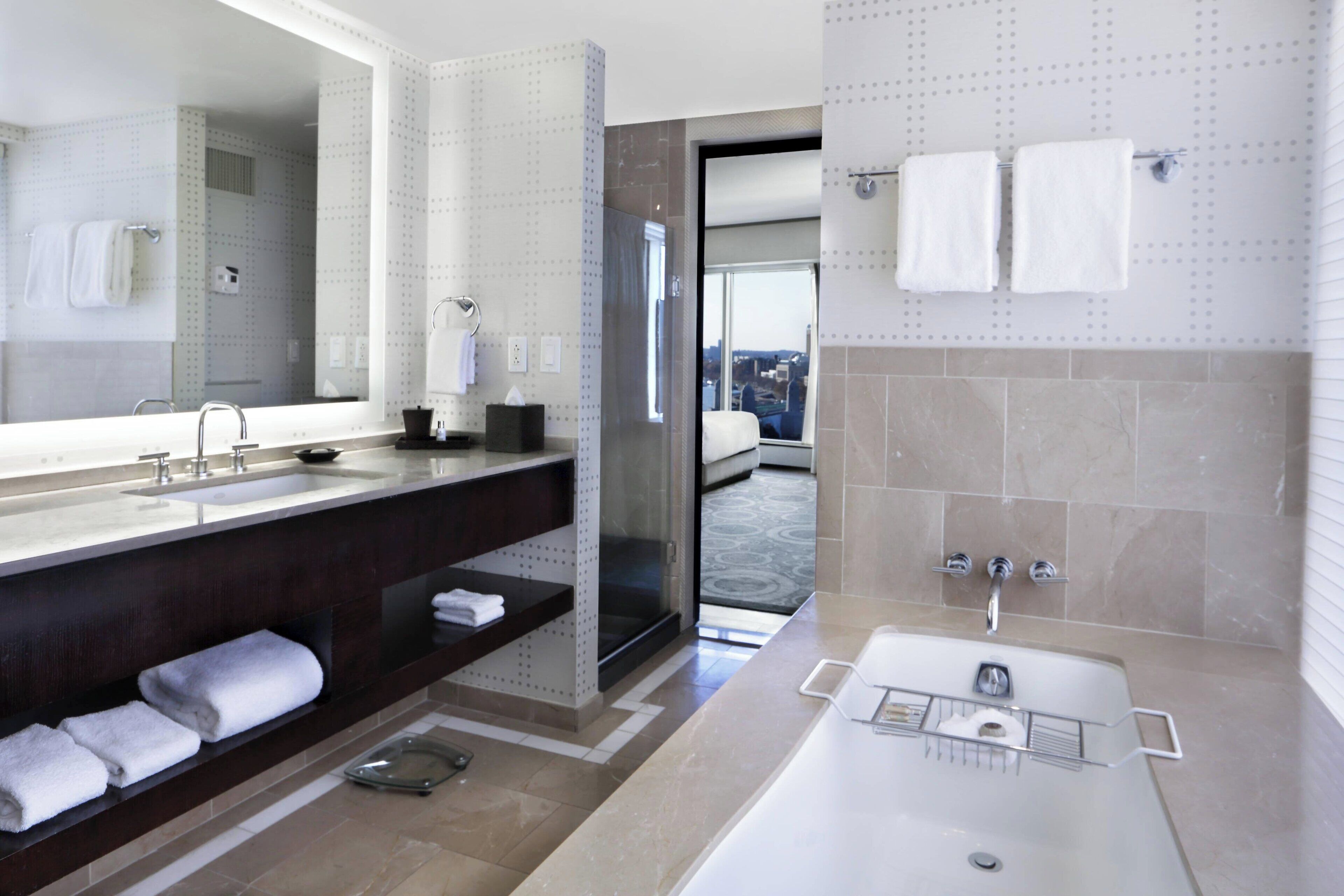 A modern bathroom with a large mirror, a bathtub, a shower, and a sink. There are towels on the counter and shelves.