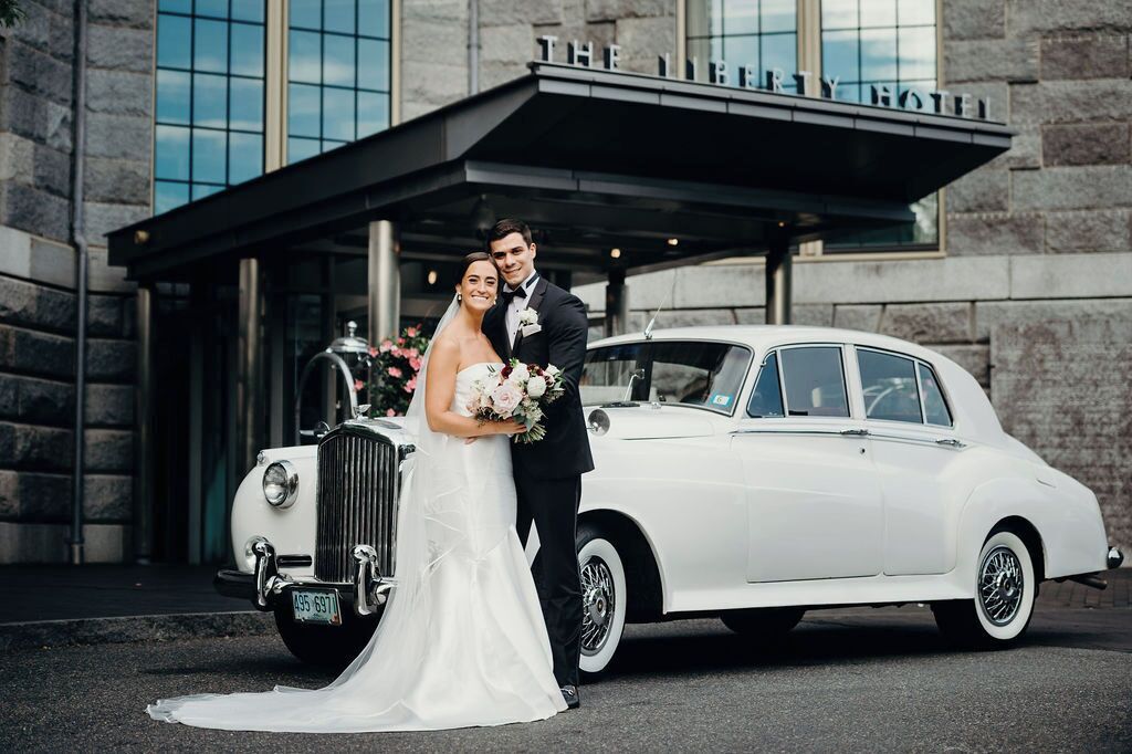 Una pareja vestida de novios posa junto a un coche blanco antiguo frente a un hotel con fachada de piedra y un cartel que dice "THE LIBRARY HOTEL".