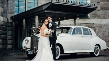 Una pareja vestida de novios posa junto a un coche blanco antiguo frente a un hotel con fachada de piedra y un cartel que dice "THE LIBRARY HOTEL".