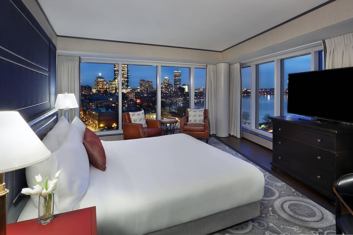 Habitación de hotel con una cama grande, televisión, cómoda y vistas a un horizonte urbano al atardecer.