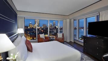 Quarto de hotel com cama grande, televisão, cômoda e vista para o horizonte urbano ao entardecer.