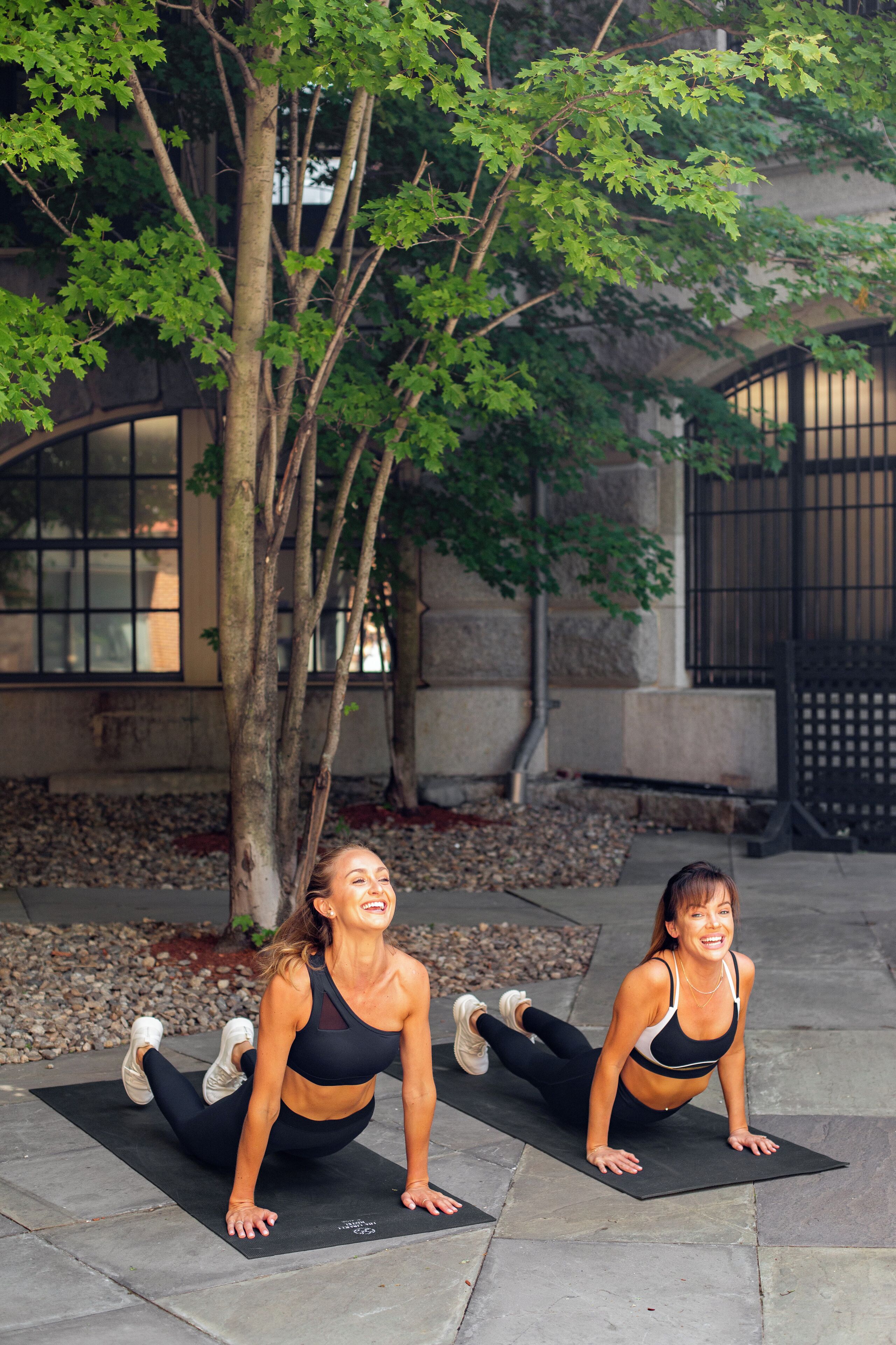 Two individuals performing a workout on a mat outdoors with trees and a building in the background.
