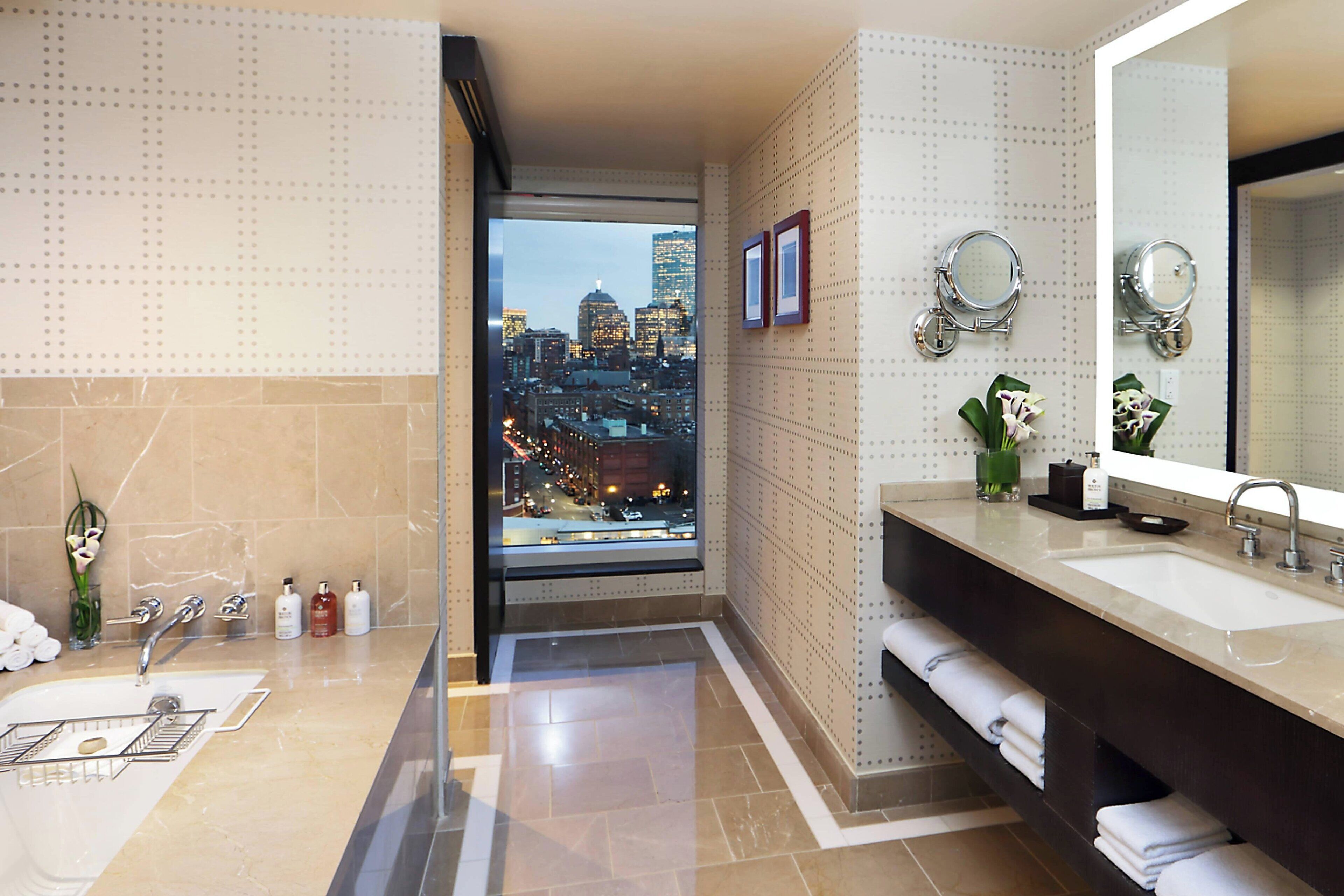 A modern bathroom with dual sinks, a large mirror, and a view of a cityscape through a large window.