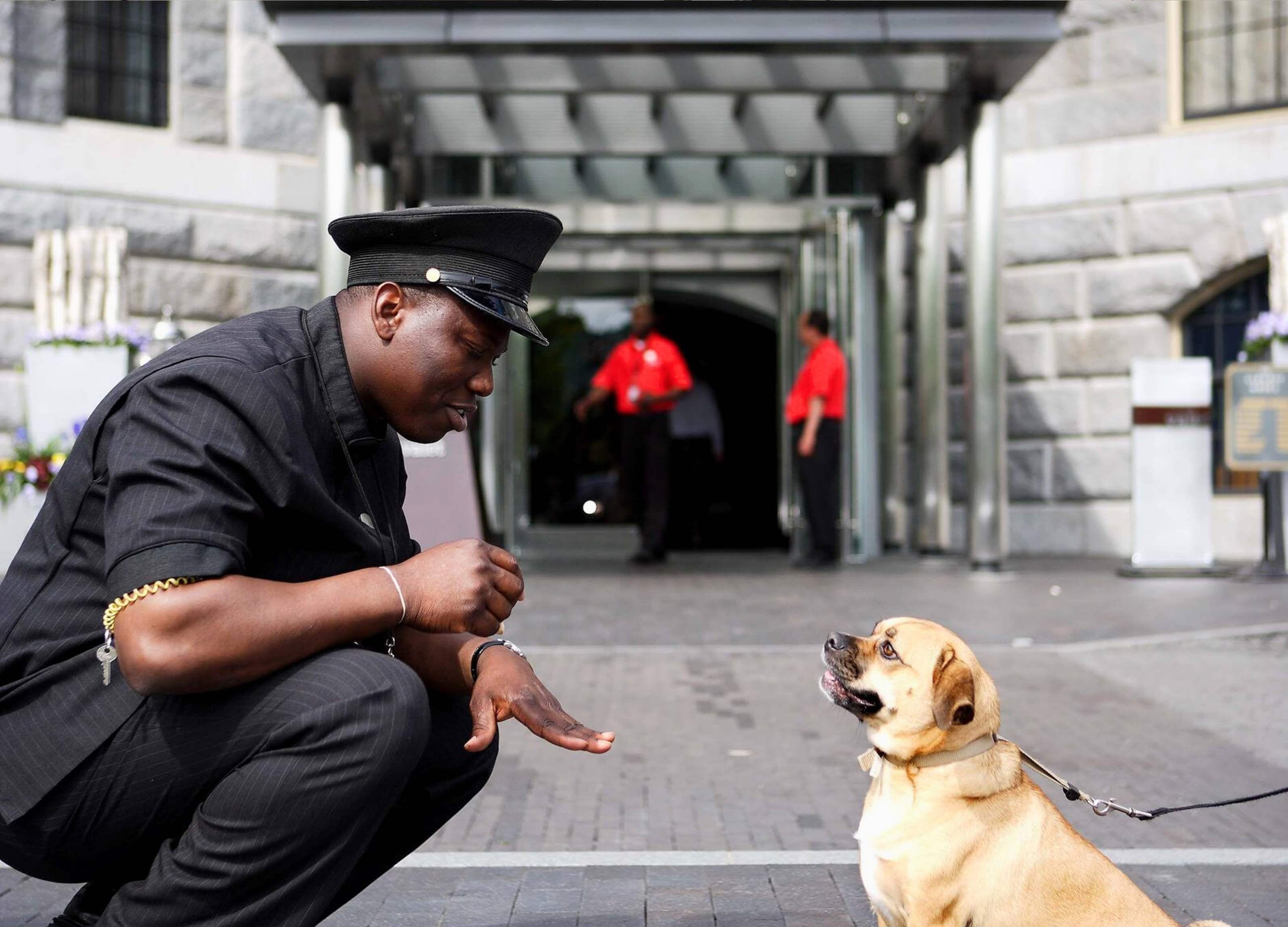 A man in uniform crouching next to a dog in an outdoor area with a building entrance in the background.