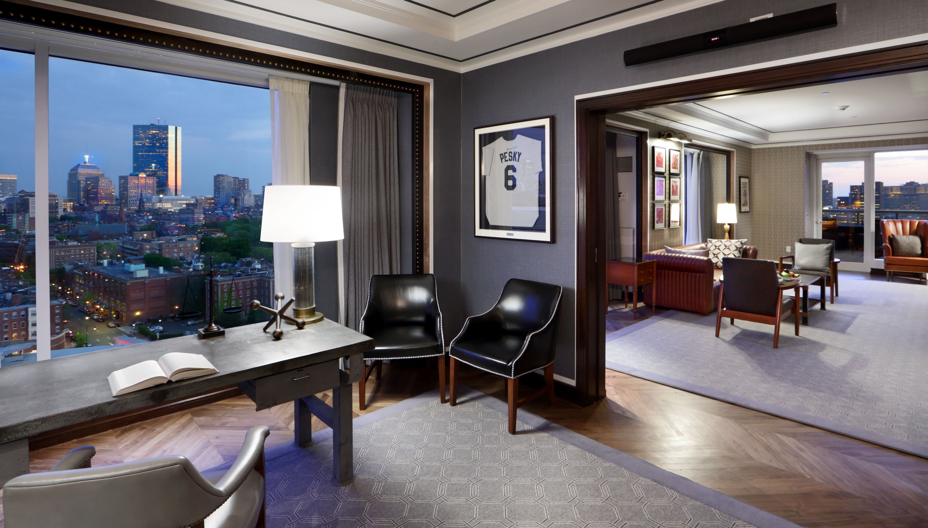 Une chambre d’hôtel moderne avec vue sur la ville, un bureau avec une lampe, deux fauteuils en cuir, un coin repas et un maillot encadré accroché au mur.