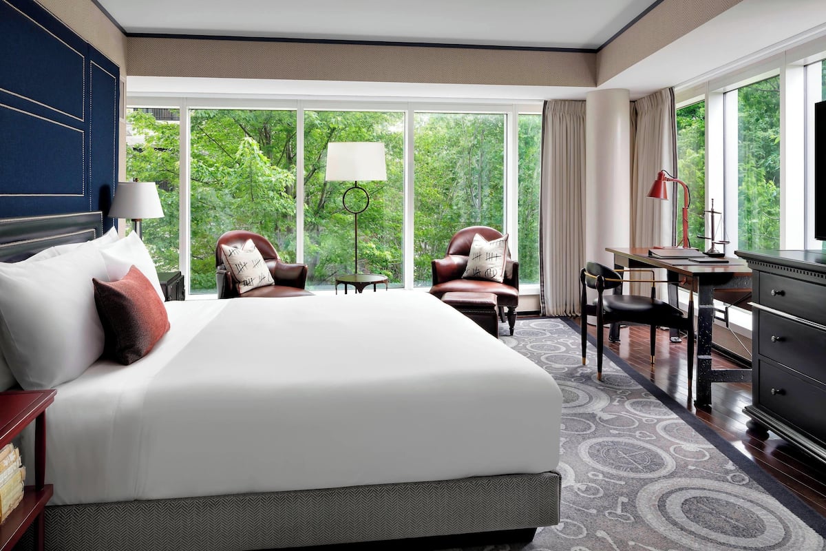 Habitación de hotel con una cama grande, dos sillones, un escritorio con lámpara y vistas a zonas verdes a través de amplios ventanales.