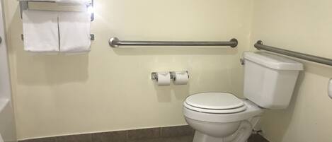 A bathroom with a toilet, a towel rack with folded towels, a handrail, and a trash bin.