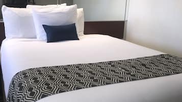 A hotel room with a large bed, white bedding, a patterned throw blanket, and a flat-screen TV mounted on the wall.