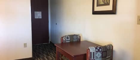 A room with a wooden table and two patterned chairs, a framed artwork on the wall, and a door with an emergency escape plan notice.