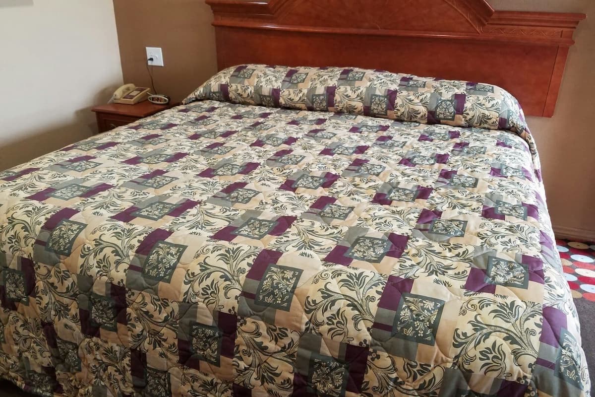 A bed with a patterned quilt, a wooden headboard, a bedside table with a telephone, and a wall socket.