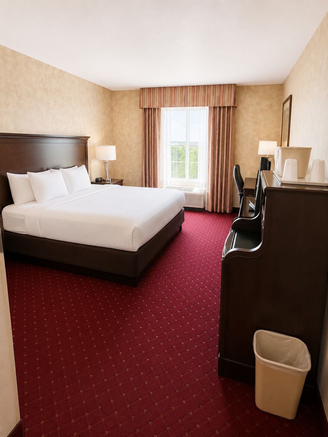A hotel room with a large bed, a desk with a chair, a waste bin, and a window with curtains.