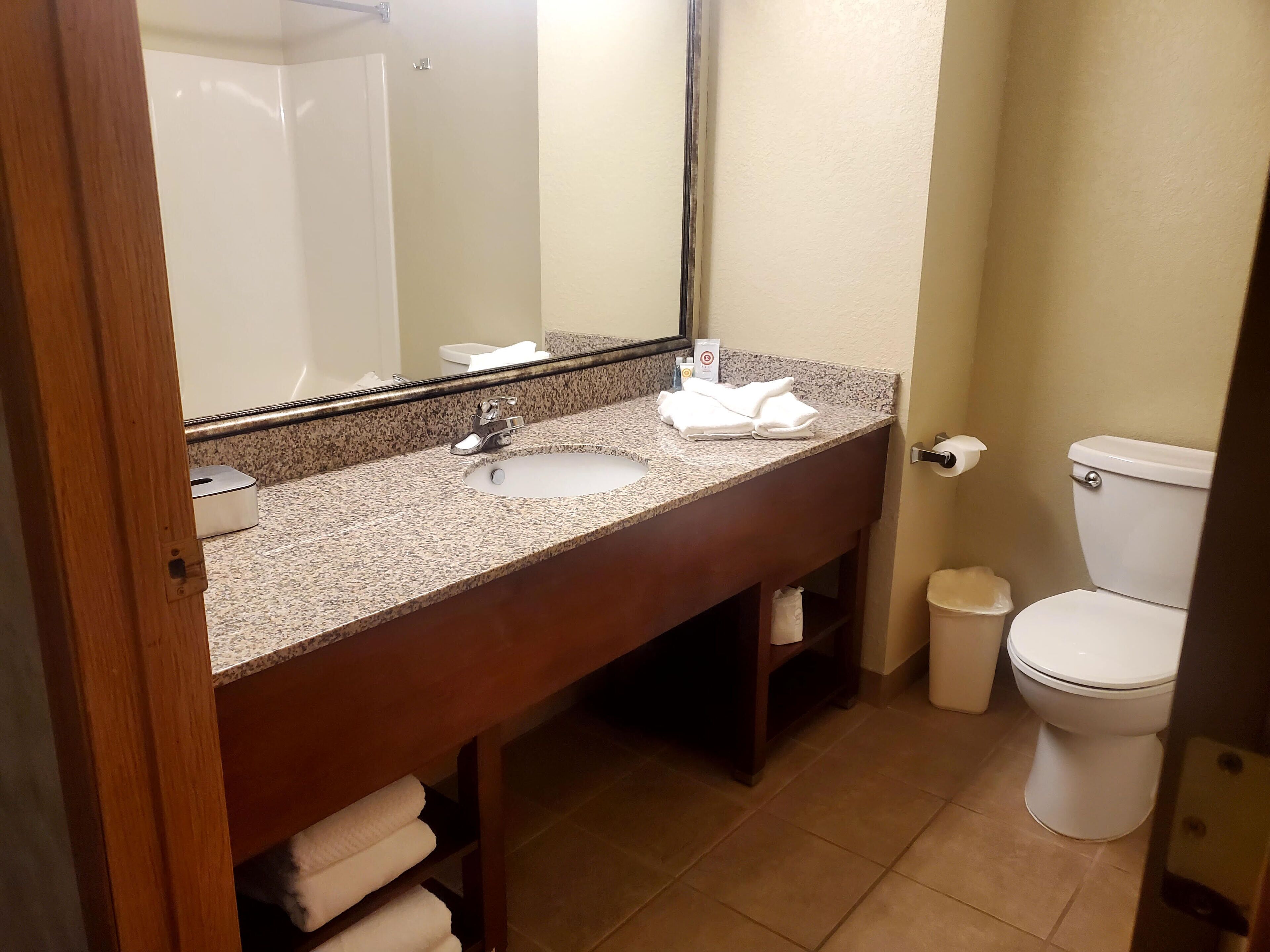 A bathroom with a granite countertop sink, a mirror, a toilet, and a trash bin.