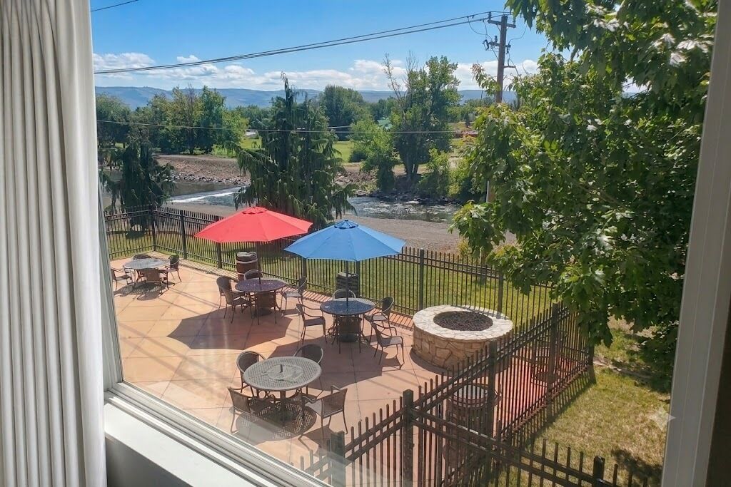 A patio area with tables and chairs, a stone fire pit, and a view of a river and mountains.