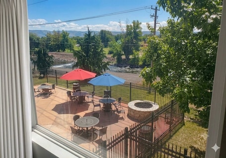 A patio with tables and chairs, a stone fire pit, and a view of a river and mountains.