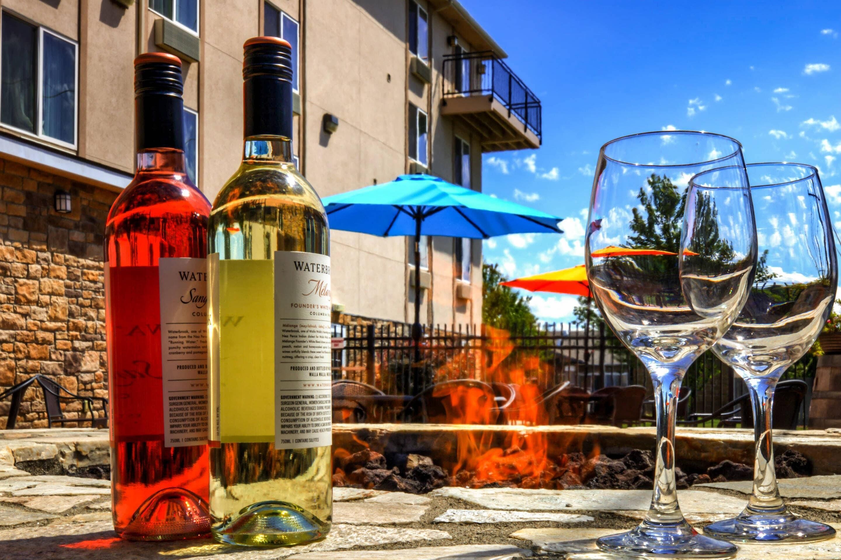 Deux bouteilles de vin et deux verres à vin sont posés sur une table à l’extérieur, avec un feu en arrière-plan.