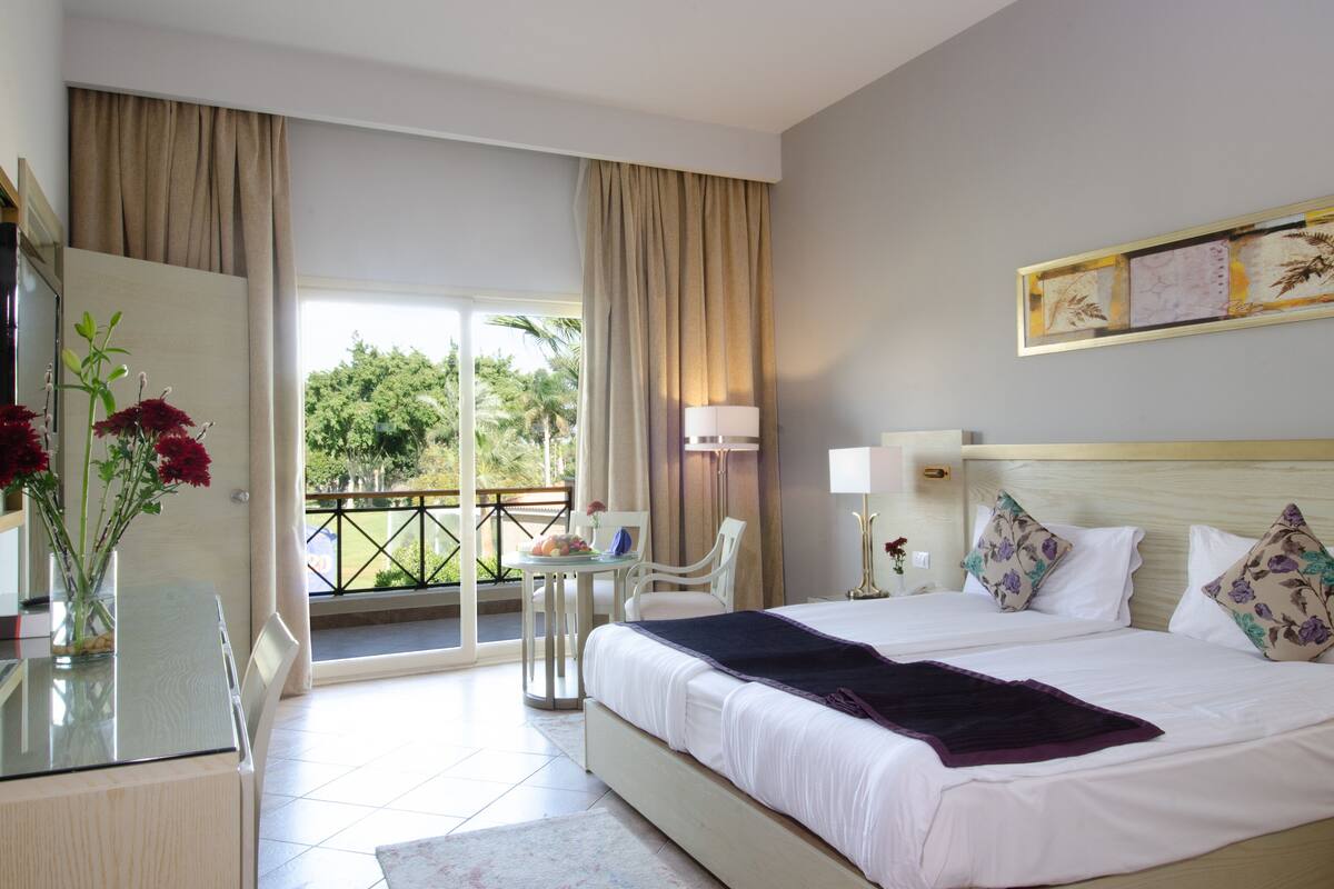 A hotel room with a bed, a desk, a chair, a balcony with a view of trees, and a painting on the wall.