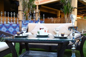 Une table à plateau en verre, sur laquelle sont disposés un plateau de thé et une théière, est placée sur une terrasse meublée de mobilier en osier, avec un décor à motifs en arrière-plan.