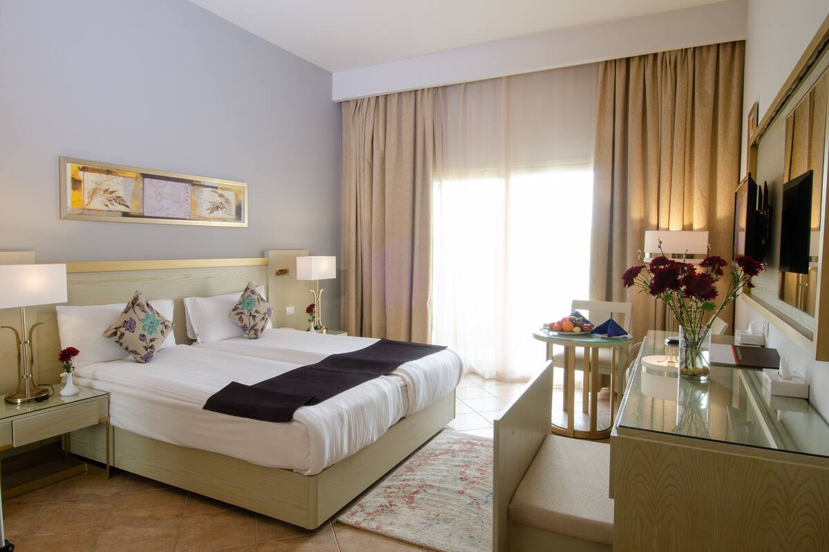 A hotel room with a large bed, bedside tables with lamps, a desk with a chair, a vase with flowers, and a large window with curtains.