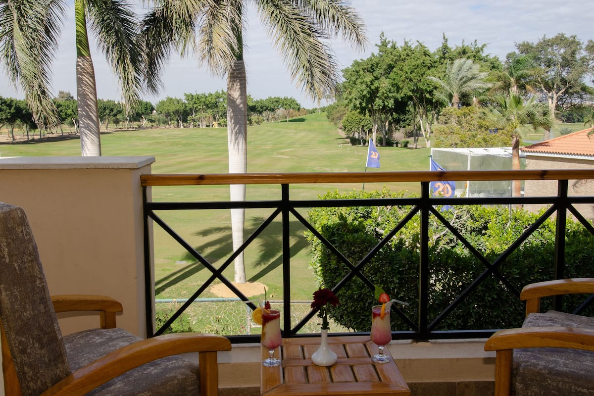 A balcony with two wooden chairs, a small table with two drinks, and a view of a golf course.