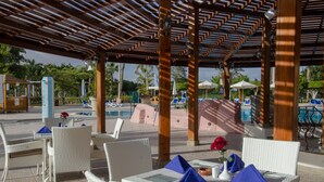 An outdoor seating area with wicker furniture and umbrellas, overlooking a pool and palm trees. - Swiss Inn Pyramids Golf Resort (6th of October City)