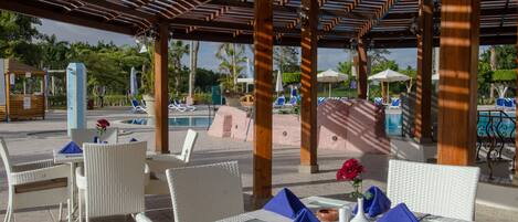 An outdoor seating area with wicker furniture and umbrellas, overlooking a pool and palm trees.