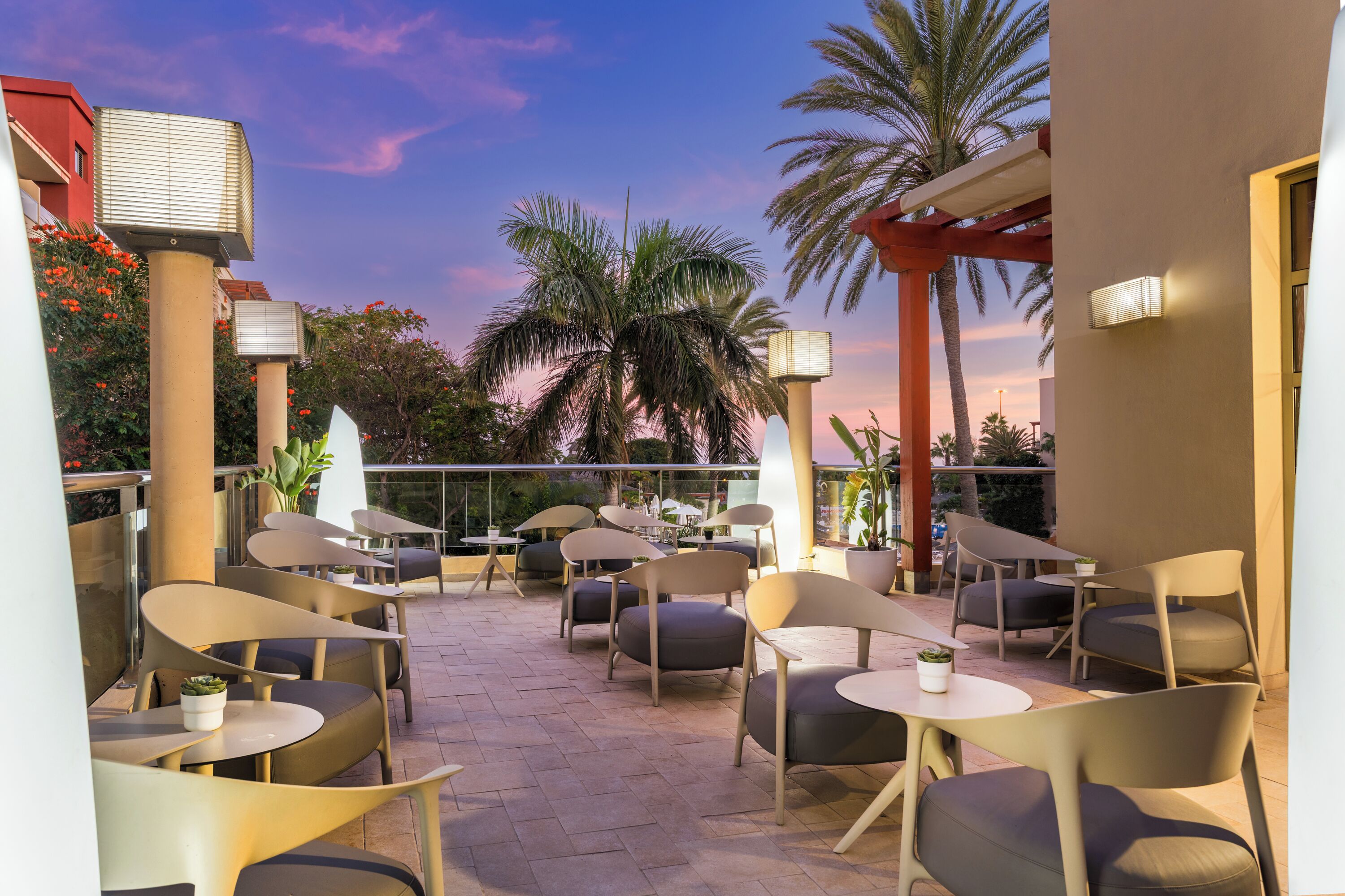 Eine Terrasse im Freien mit mehreren Tischen und Stühlen, Palmen und einem Sonnenuntergangshimmel.