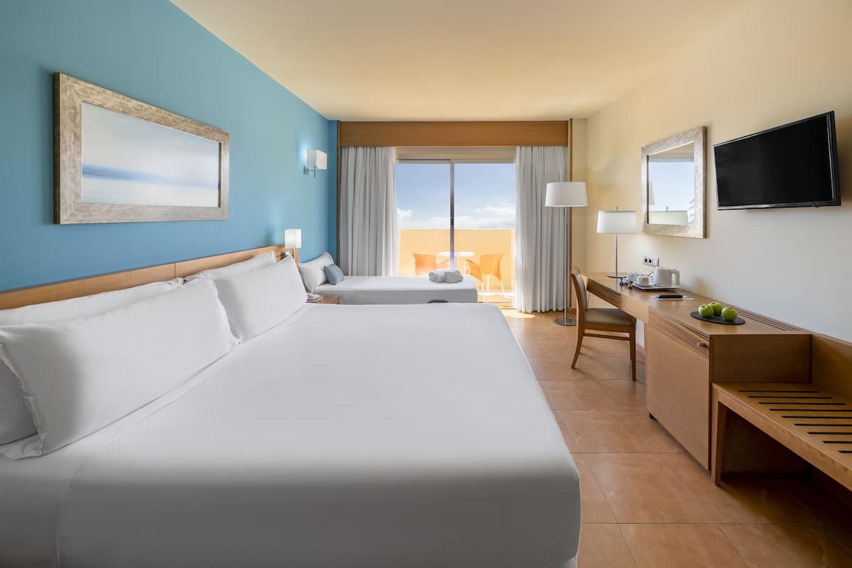 Ein Hotelzimmer mit zwei Betten, einem Schreibtisch, einem Stuhl, einem Fernseher und einem Balkon mit Tisch und Stühlen.