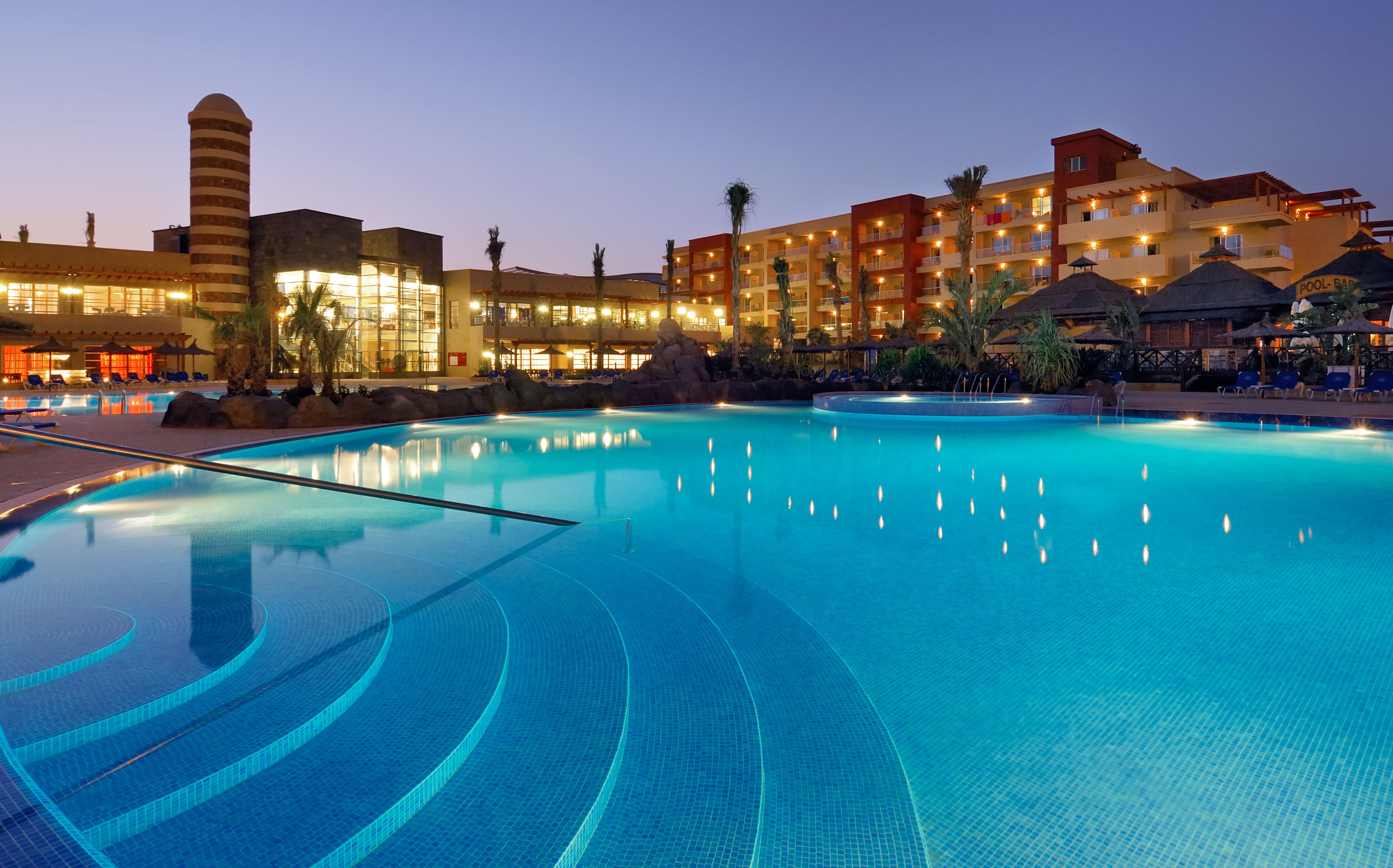 A large indoor swimming pool with clear water, surrounded by a hotel with multiple balconies and a rock formation.