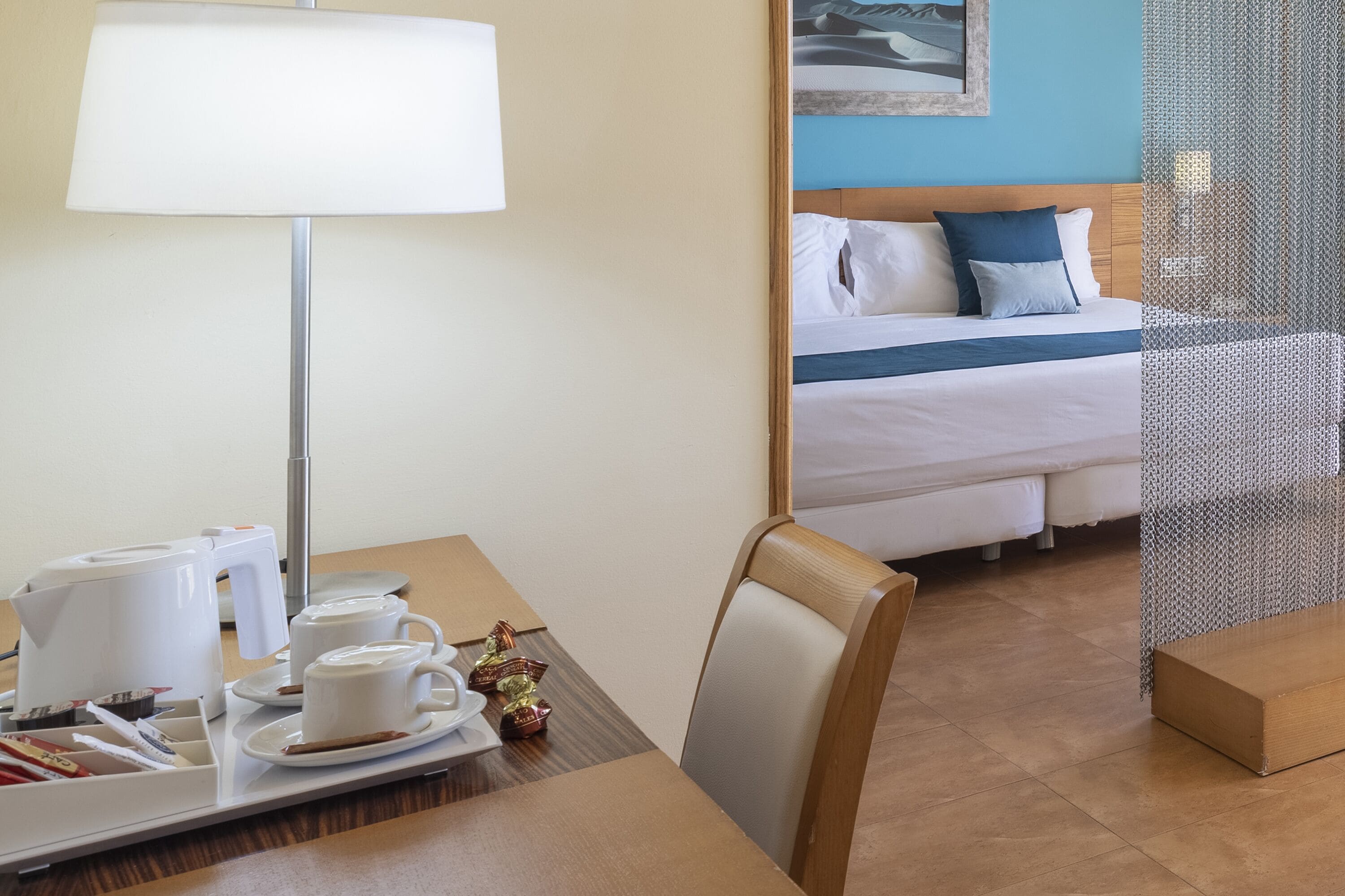 A hotel room with a bed, a bedside table with a lamp, a tray with cups and a teapot, and a breakfast setup.
