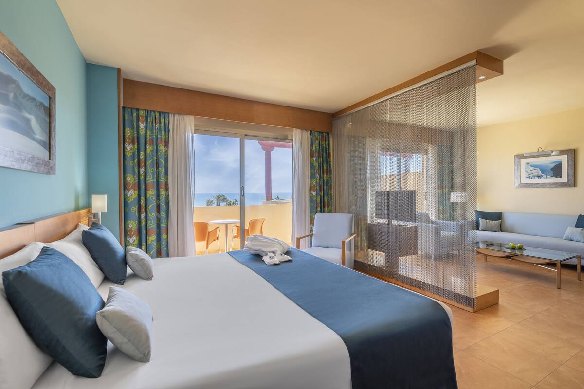 Ein Hotelzimmer mit einem großen Bett, einem Balkon mit Aussicht, einer Sitzecke und einem Fernseher.
