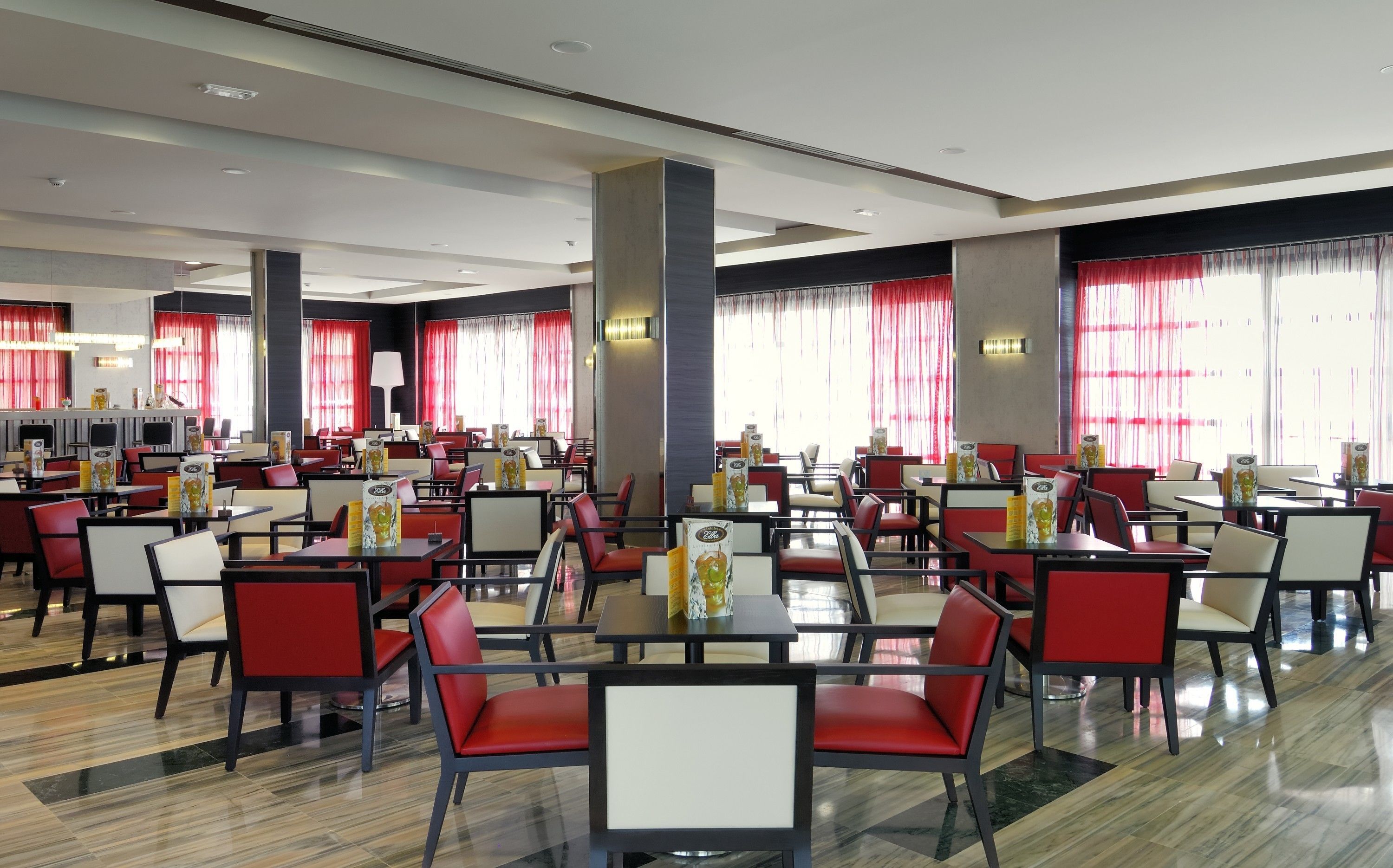 En rummelig hotelrestaurant med flere borde og stole, store vinduer med røde gardiner og et moderne loftsdesign.