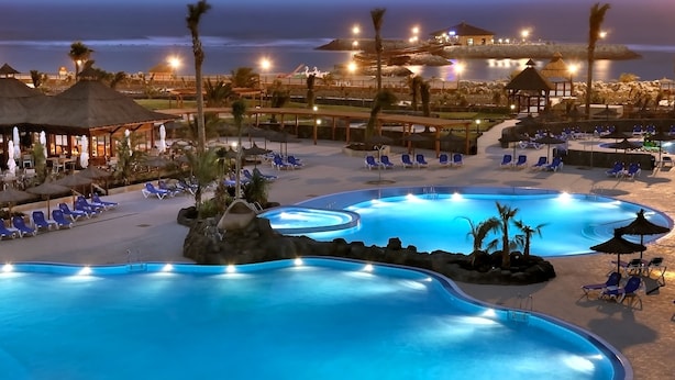 Zona de piscina en un complejo turístico con varias piscinas, sillones de descanso, sombrillas y vistas al océano.