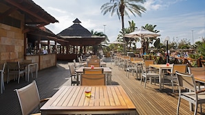 Un espacio de comedor al aire libre con mesas y sillas de madera, una zona de bar y una estructura con techo de paja.