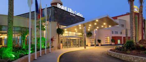 Fachada de hotel de diseño moderno, con estructura de vidrio, zona de acceso cubierta y un camino bien iluminado que lleva a la entrada.