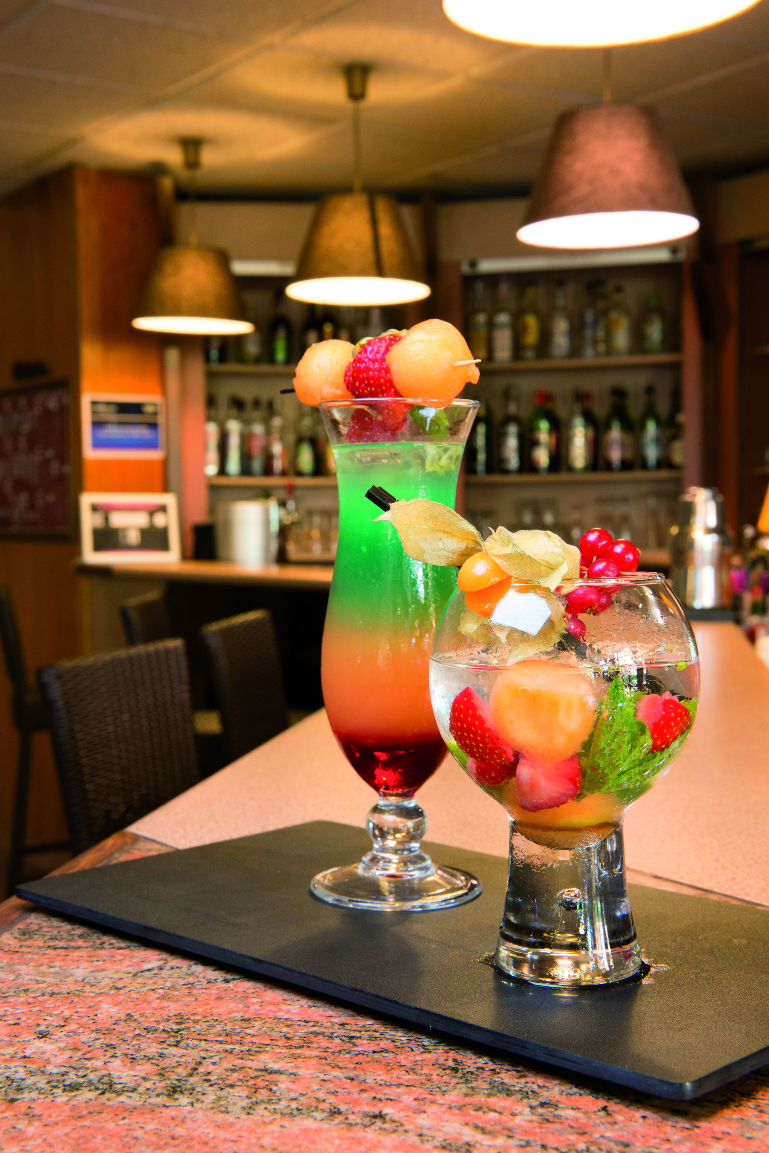 Dois cocktails sobre uma mesa, com um bar ao fundo.
