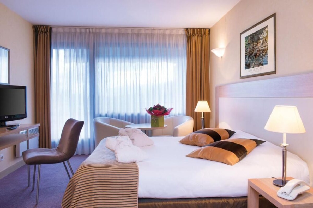 A hotel room with a bed, a desk with a television, a chair, a table with a vase of flowers, and a lamp.