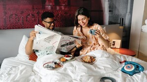 Una pareja sentada en una cama con un periódico, una taza y un teléfono.