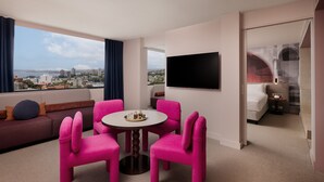 Habitación de hotel con vista a la ciudad, televisor de pantalla plana, mesa de comedor con sillas rosas, sofá y cama.