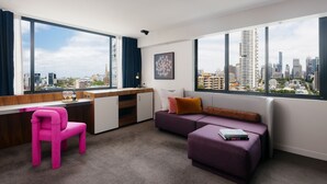 Una habitación moderna con vistas a la ciudad, un escritorio, una silla rosa, un sofá morado y un radiador.