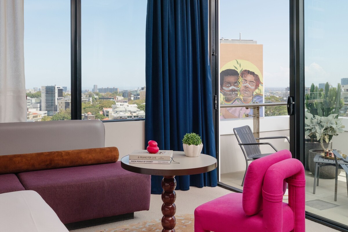 A modern hotel room with a large window, a sofa, a chair, a small table with a plant, and a poster on the wall.