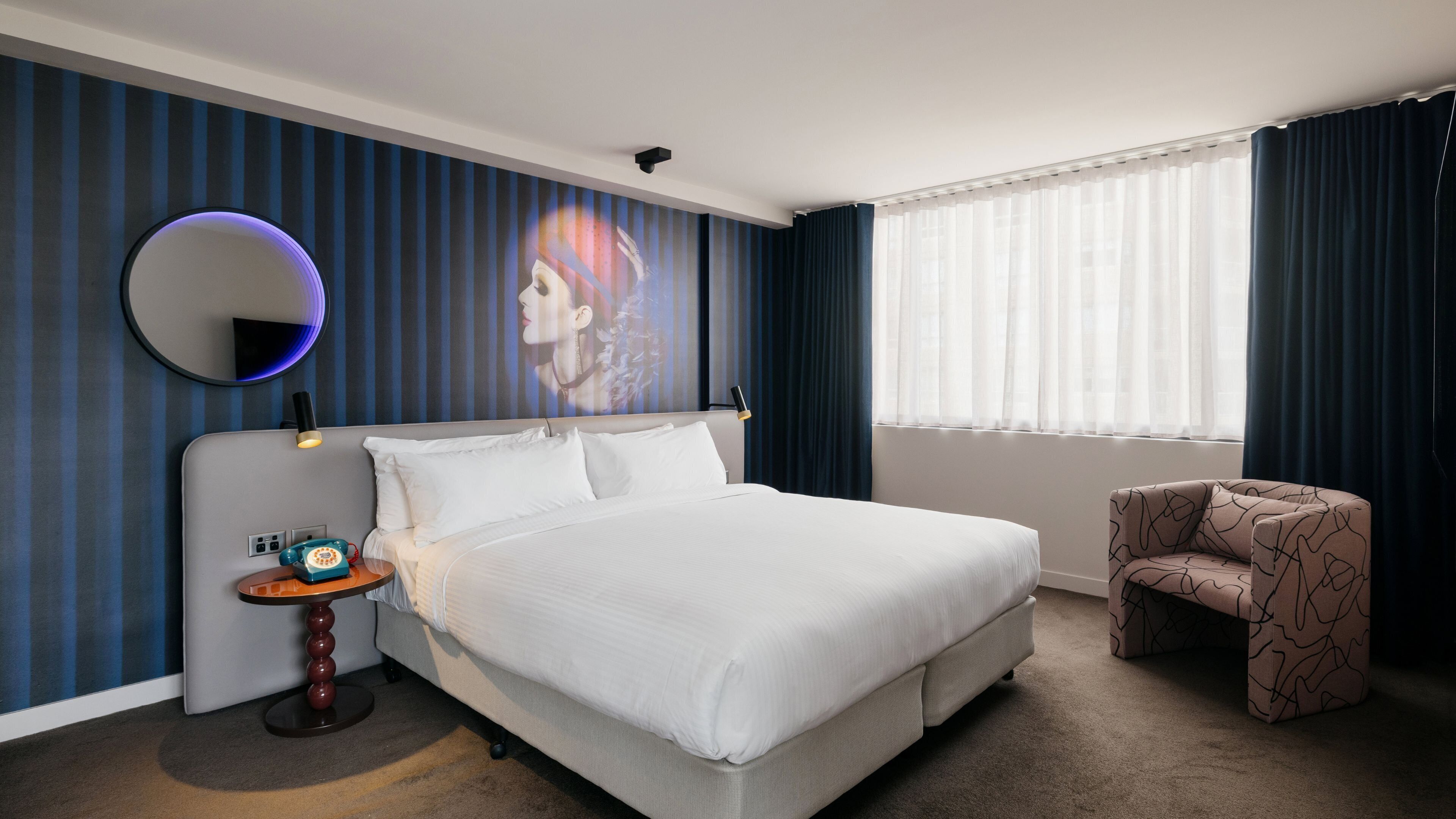 A hotel room with a large bed, a round mirror, a small table with a telephone, and a patterned armchair.